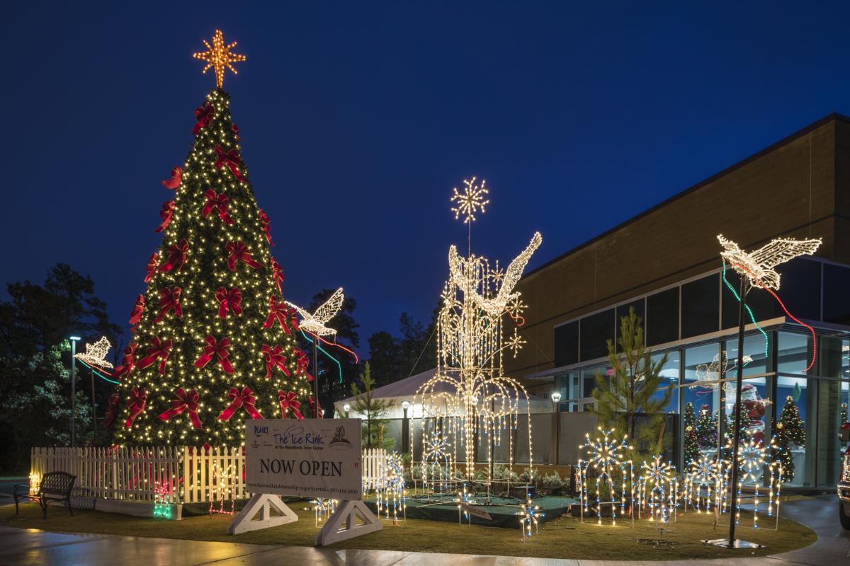 The Woodlands Ice Rink exterior. On the left is a giant Christmas tree decked with golden lights and large red ribbons. Various light displays (shaped like doves, a fountain, snowflakes, and more) line the walkway to the building. A sign reads "The Ice Rink at The Woodlands Town Center | NOW OPEN."