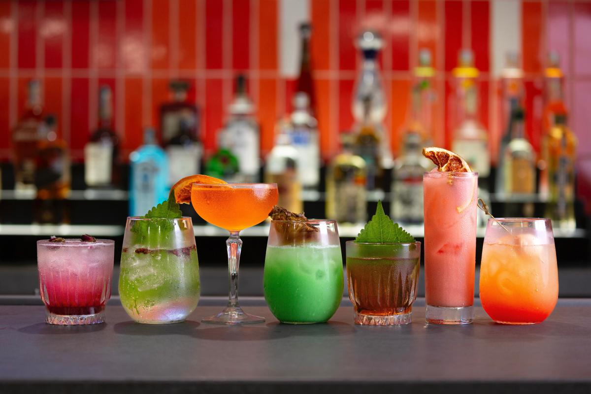 The camera faces the bar (from the height of the bar), which is topped with seven glasses of various shapes and sizes, each filled with a unique and colorful cocktail. The first and fifth glasses are short and squatty, one filled with a bright pink drink topped with ice while the other is burgundy with a large herbal leaf peeking out the top. The second, fourth, and seventh glasses are medium-height and teardrop shaped: one containing a clear liquid with a decorative leaf, another features a bright minty green liquid with a decorative mushroom (?), and another with a bright sunset-colored drink and a toothpick. The third glass in the row is a martini glass filled with a blood orange-colored drink and slice. The sixth glass is tall and thin with a light pink drink and blood orange slice.