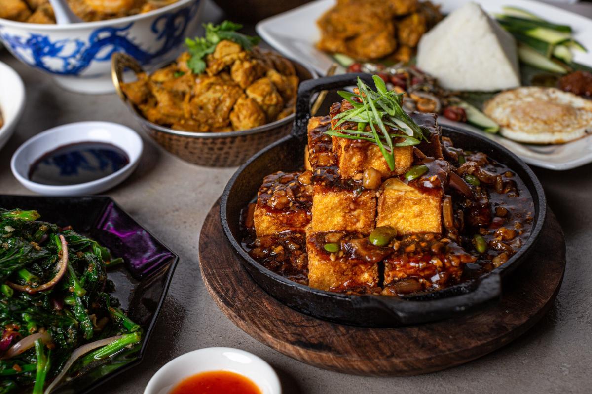 The camera zooms in on a table piled high with various Asian dishes. The one closest to the viewer appears to be a fried tofu skillet (white cubes stacked like a pyramid, doused in a nutty, orange-colored sauce, and topped with herbs). Surrounding this piece are various other entrees and appetizers. One looks to be fried chicken, another, steamed veggies. A third has fried chicken, steamed veggies, a pyramid of rice, and what looks to be a small pancake.