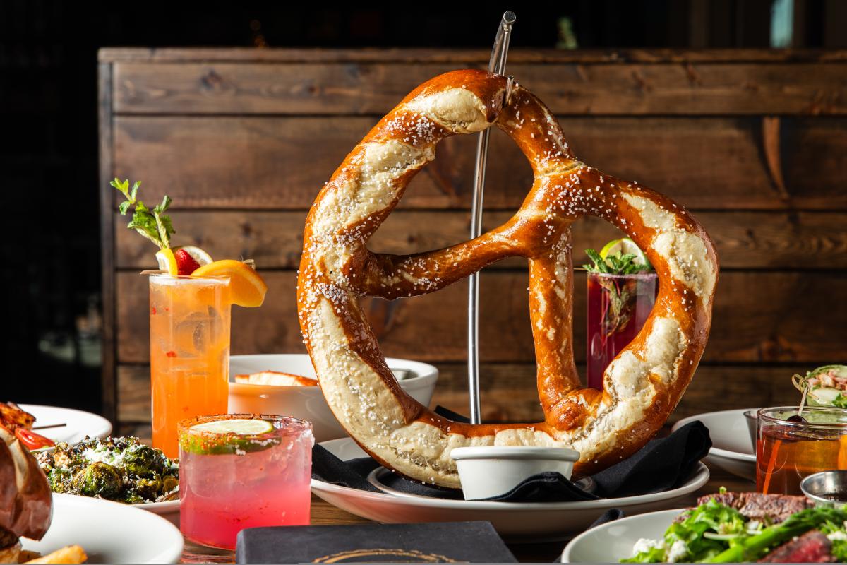 A close-up view of a giant pretzel hanging from a metal hook. Around it are happy hour drinks, including a bourbon and two cocktails (one orange, one pink) topped with fruit slices.