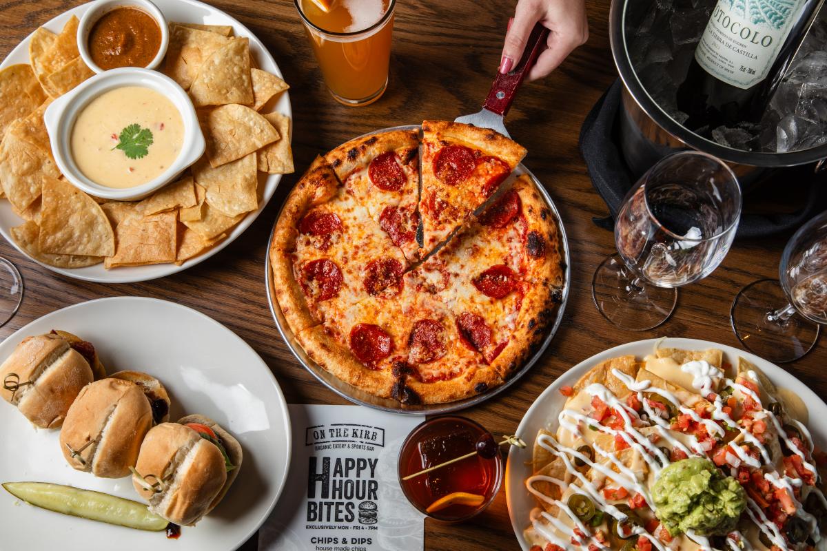 An overhead view of food from On the Kirb. A small pepperoni pizza is in the center with a woman's hand taking a slice. Around it are a bucket of ice with a wine bottle inside, a plate of loaded nachos, a burboun, a plate of three sliders, and a plate of chips with queso and salsa.