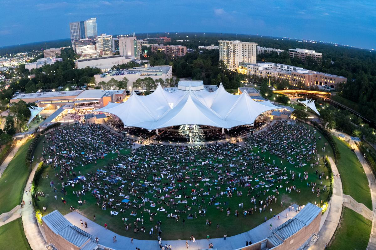 An aerial view of The Cynthia Woods Mitchell Pavilion at dusk. The seats and lawn are packed full of people for the Parker McCollumn concert. Nearby hotel and office lights are just starting to light up for the evening.