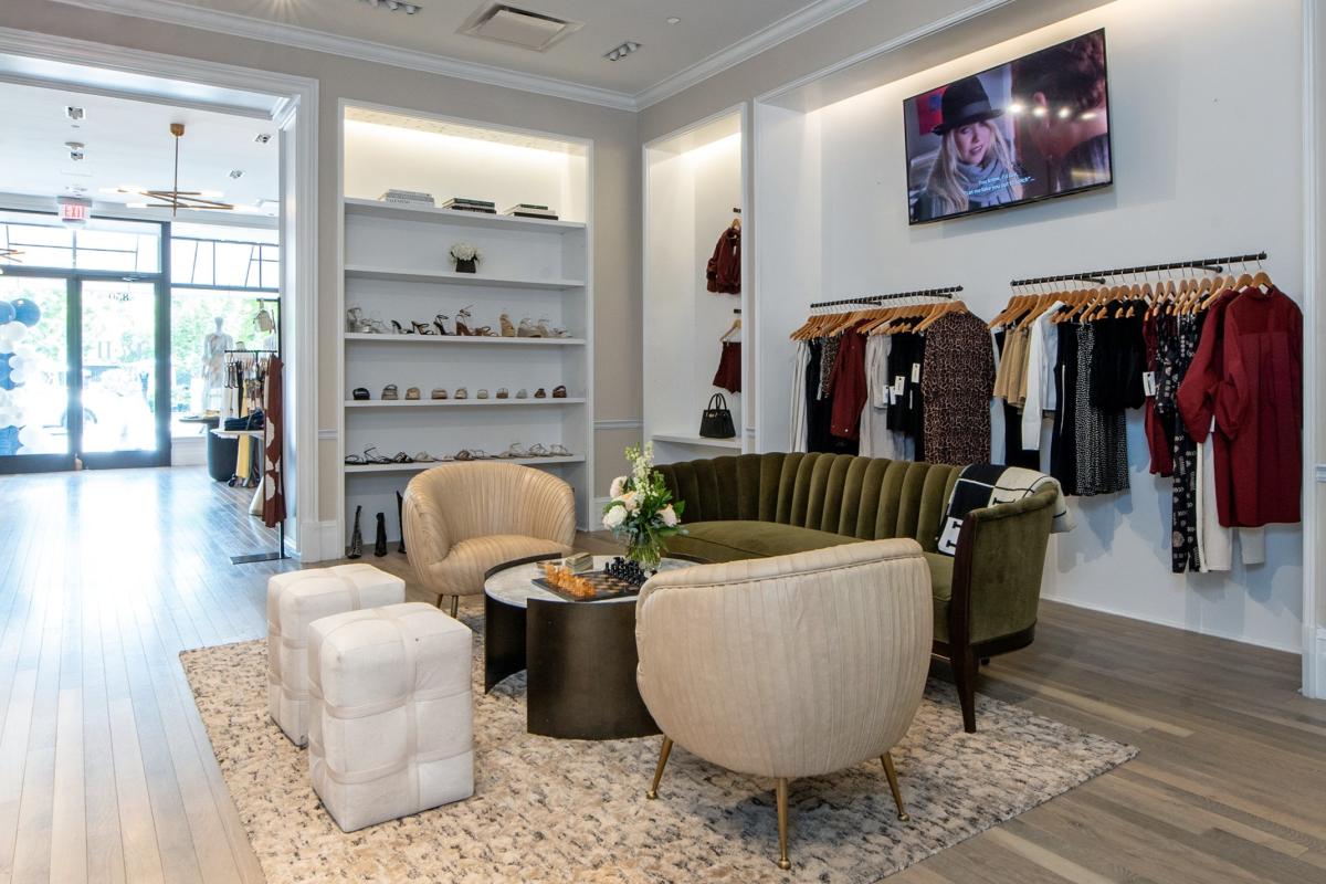 The camera overlooks the interior of Pell 1990 at Market Street. Closest to the camera is a small sitting area with two cream-colored satin chairs, a plush olive-green couch, and a round, white, marble coffee table. Behind it is a display of trendy women's clothes and shoes.