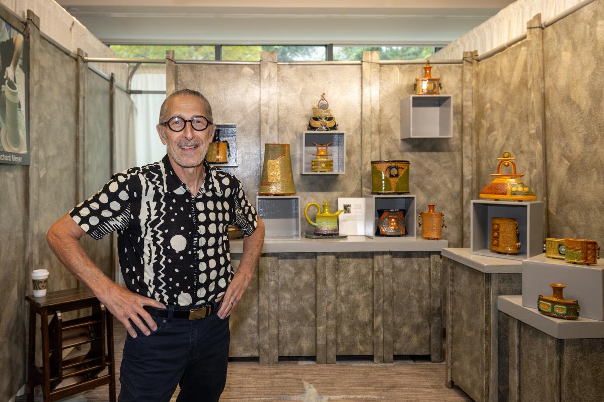 A man, 60s, smiles at the camera. He's wearing round, thick-rimmed glasses and a black shirt with large white dots and scribbles. Behind him is a booth displaying his ceramic artwork.