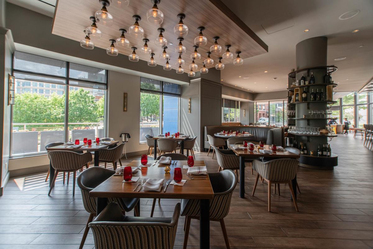 Tables for four are seated beside a floor-to-ceiling window overlooking The Waterway. The tables are set with four wine glasses, water glasses, and red-and-white cloth napkins placed atop a simple white plate. The chairs are small but plush. The lighting is dim, but the room is lit with daylight.