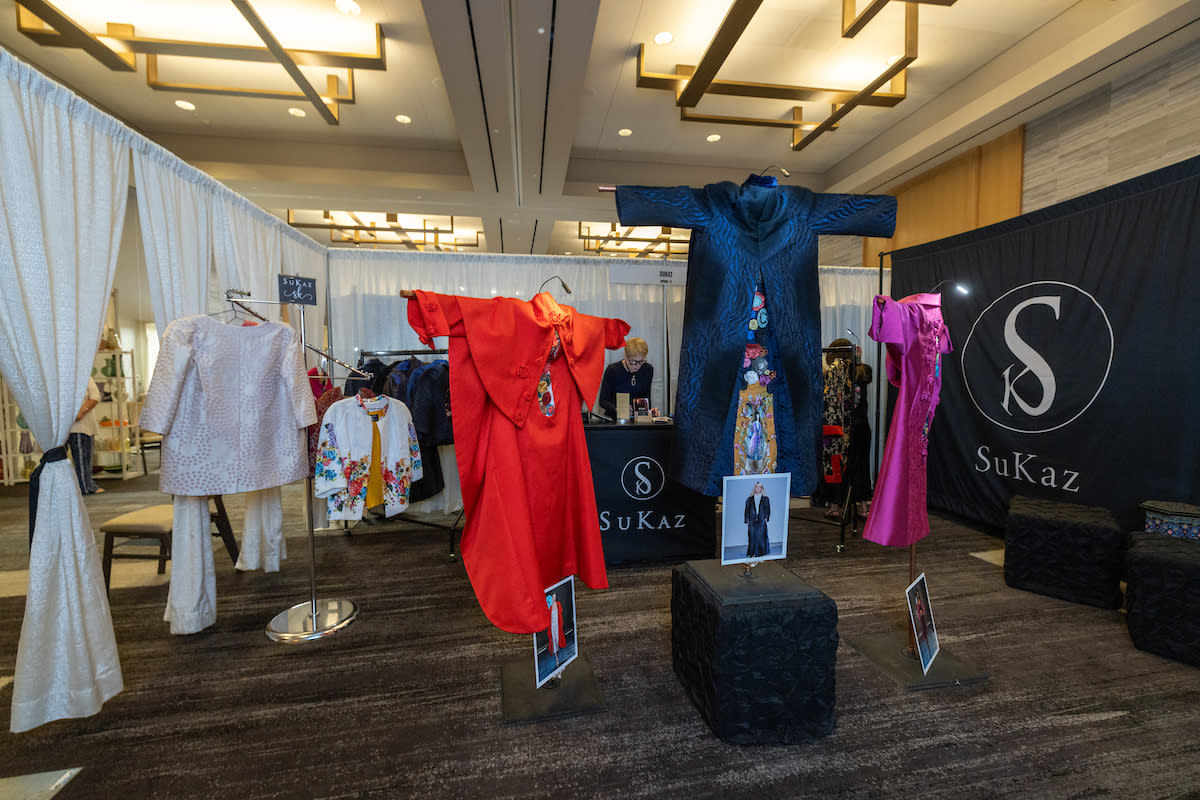 A art booth sectioned off by fancy white curtains on three sides and a black banner emblazoned with the "SuKaz" logo. Three East Asian-inspired clothing designs are on display in a T-shaped pose: a bright red cape, a deep blue quilted robe, and a bright magenta jacket, all with colorful floral embroidery.