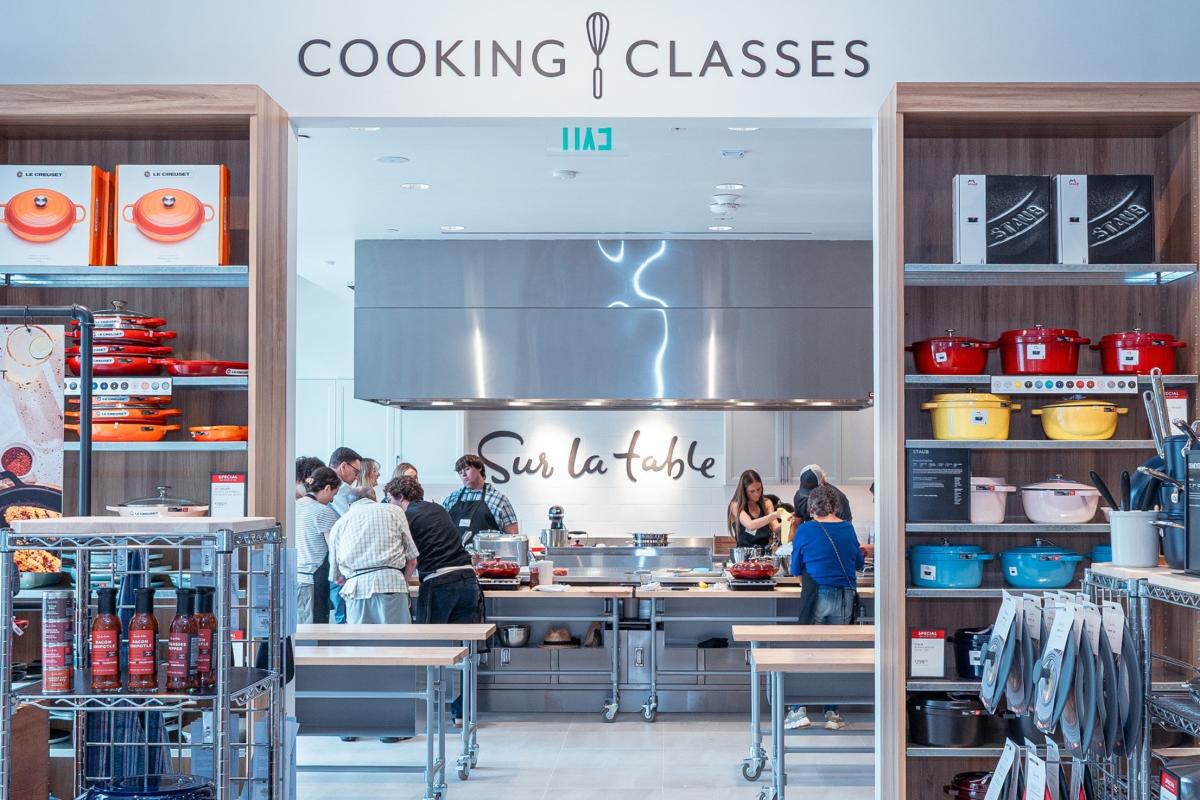 The camera looks through the kitchen supplies for sale at Sur La Table into their kitchen, which is holding a cooking class for men and women of various ages and backgrounds.