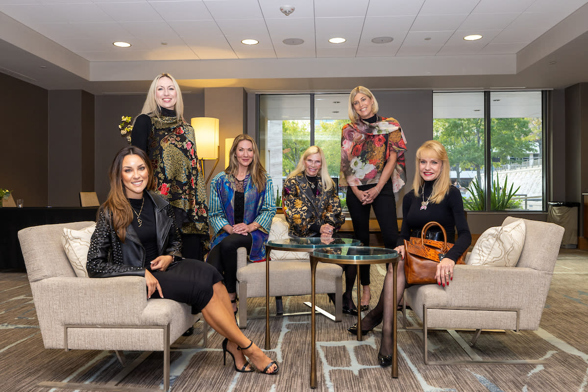 Six well-dressed women smile at the camera. Four of them are sitting on fancy white armchairs angled toward each other. The remaining two women stand between them.