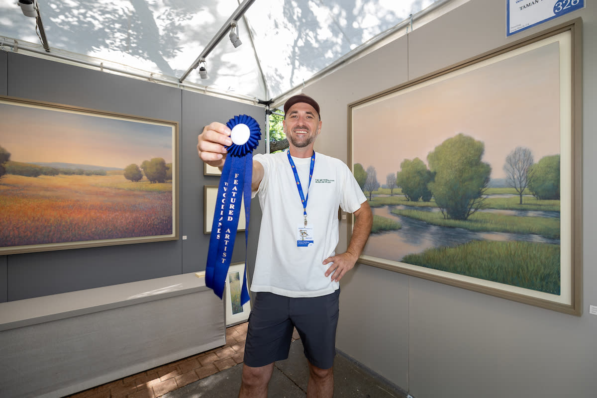 A man in a white T-shirt and gray shorts smiles at the camera. He has one hand on his hip, the other holding a blue ribbon out in front of him. Behind him are two large landscape paintings, one of a field of wildflowers and the other of wetlands.