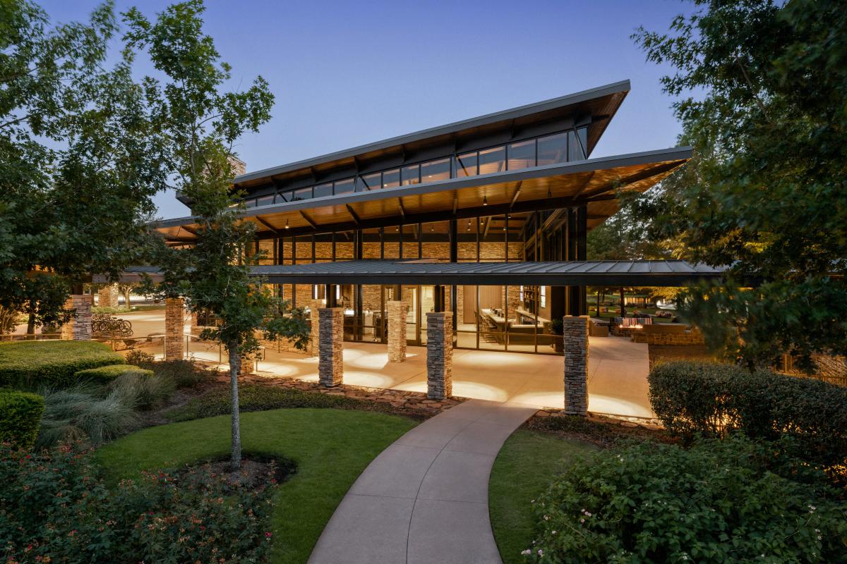 An exterior shot of The Woodlands Resort. The lobby is in a tall one-story building with a three-tiered roof, where each tier slants slightly more upward. Decorative brick columns support the first tier. The lights in the lobby and under the awning glow with soft golden light. The surrounding area is lush with neatly trimmed foliage.