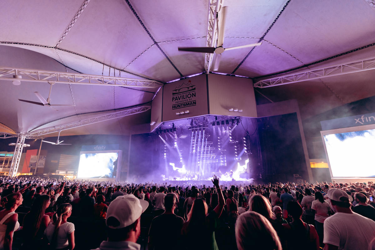 The camera faces The Pavilion stage from the right side of the crowd. Fake smoke and dozens of white and purple spotlights surround the performer (Post Malone, not clearly visible) while spectators cheer. Two big screens are on either side of the stage to show close-ups of the stage (also not clear in the photo). Above the stage is a large logo of The Pavilion and dark text that reads, "THE CYNTHIA WOODS MITCHELL PAVILION PRESENTED BY HUNTSMAN."