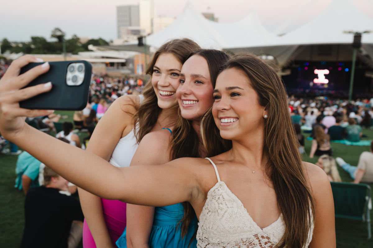 Three teen girls in colorful concert attire take a selfie on The Pavilion lawn.