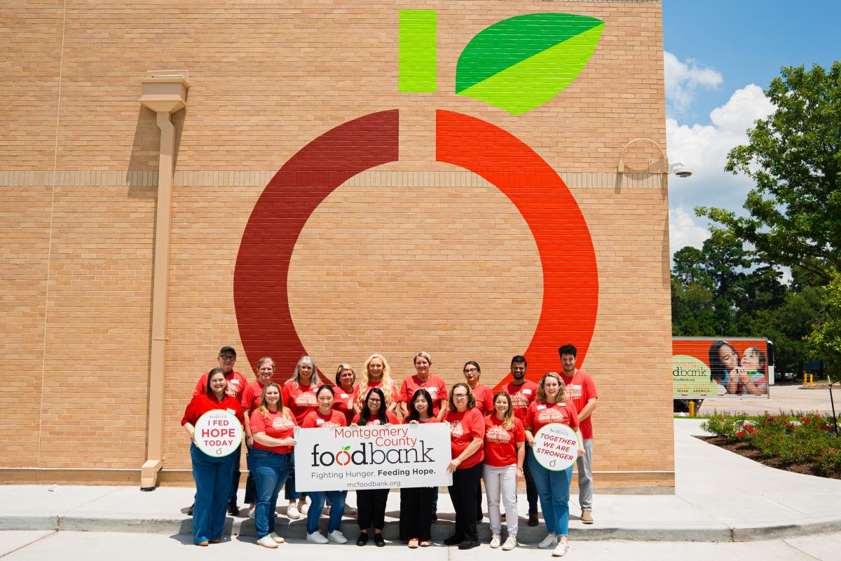 The MoCo Food Bank is a tan-brick building a mural of the food bank logo (a minimalist apple with a red circle, green stem, and green leaf). The Sales & Marketing Committees are standing in front of the mural with signs that read, "I FED HOPE TODAY," "MONTGOMERY COUNTY FOOD BANK | Fighting Hunger, Feeding Hope," and "TOGETHER WE ARE STRONGER."