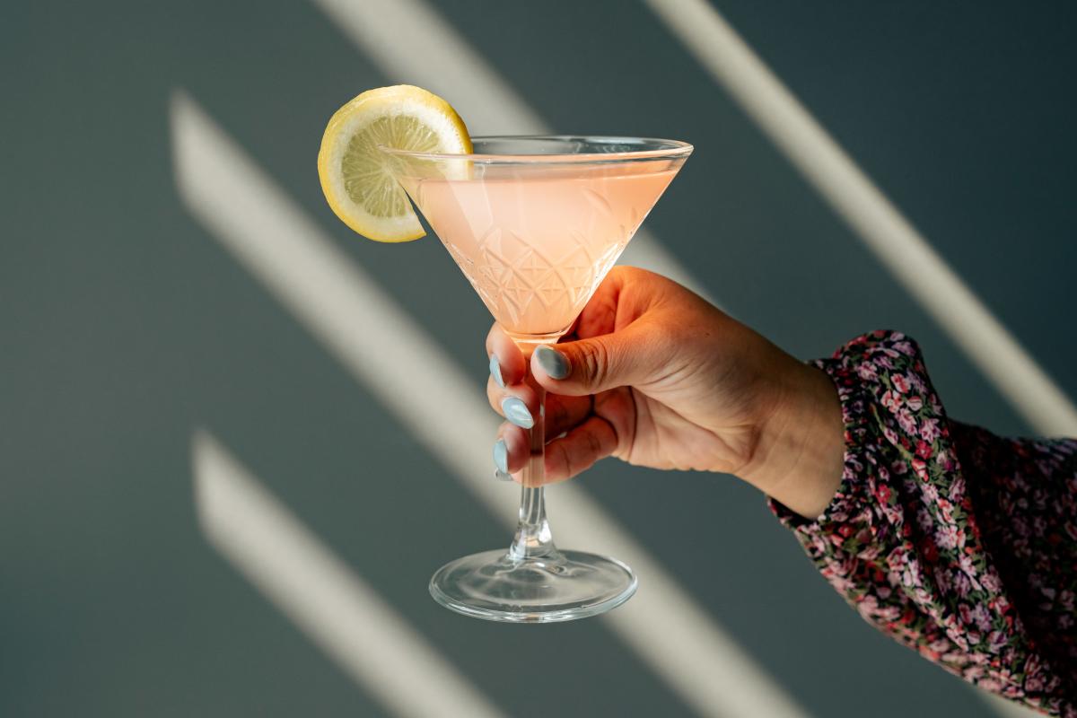 A young woman (off-screen) wearing a floral blouse holds up a light pink martini with a lemon wedge on a solid gray background. Four beams of light stream across the subjects through blinds (off-screen).