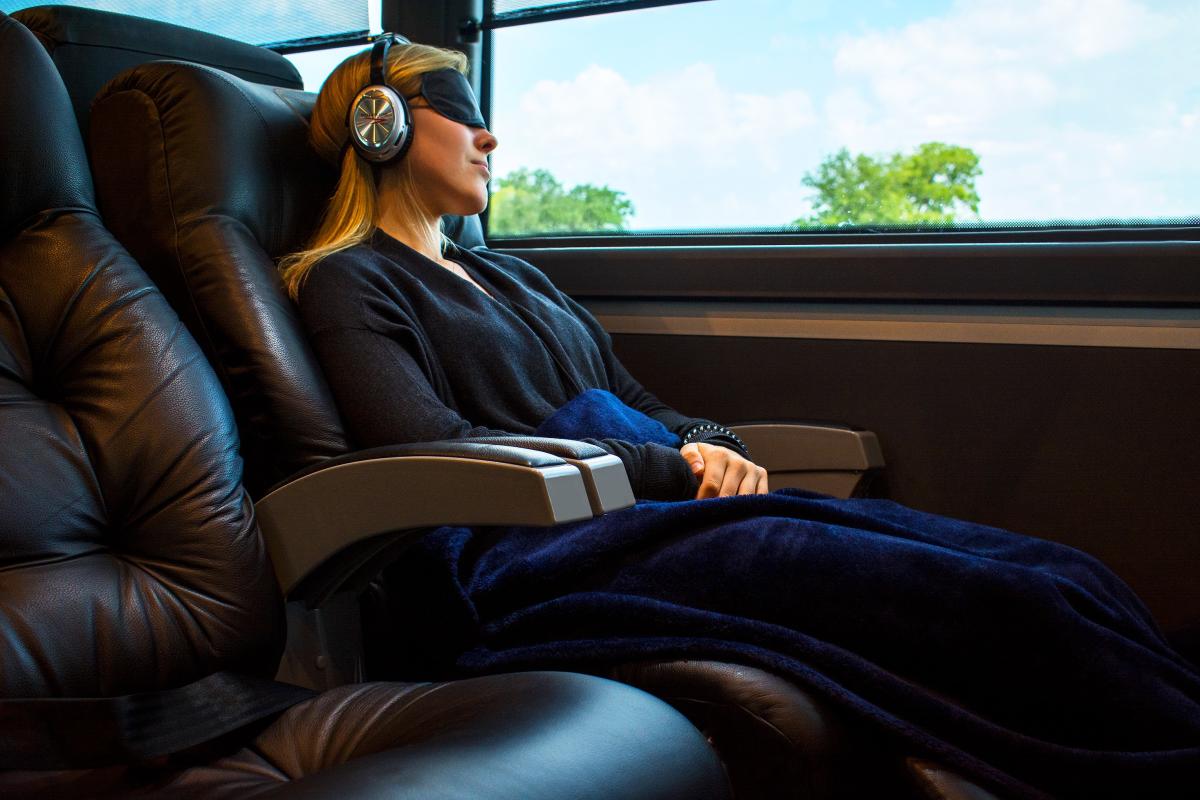 A blonde woman wears a black sleep mask and silver headphones as she reclines in a leather Vonlane bus seat.