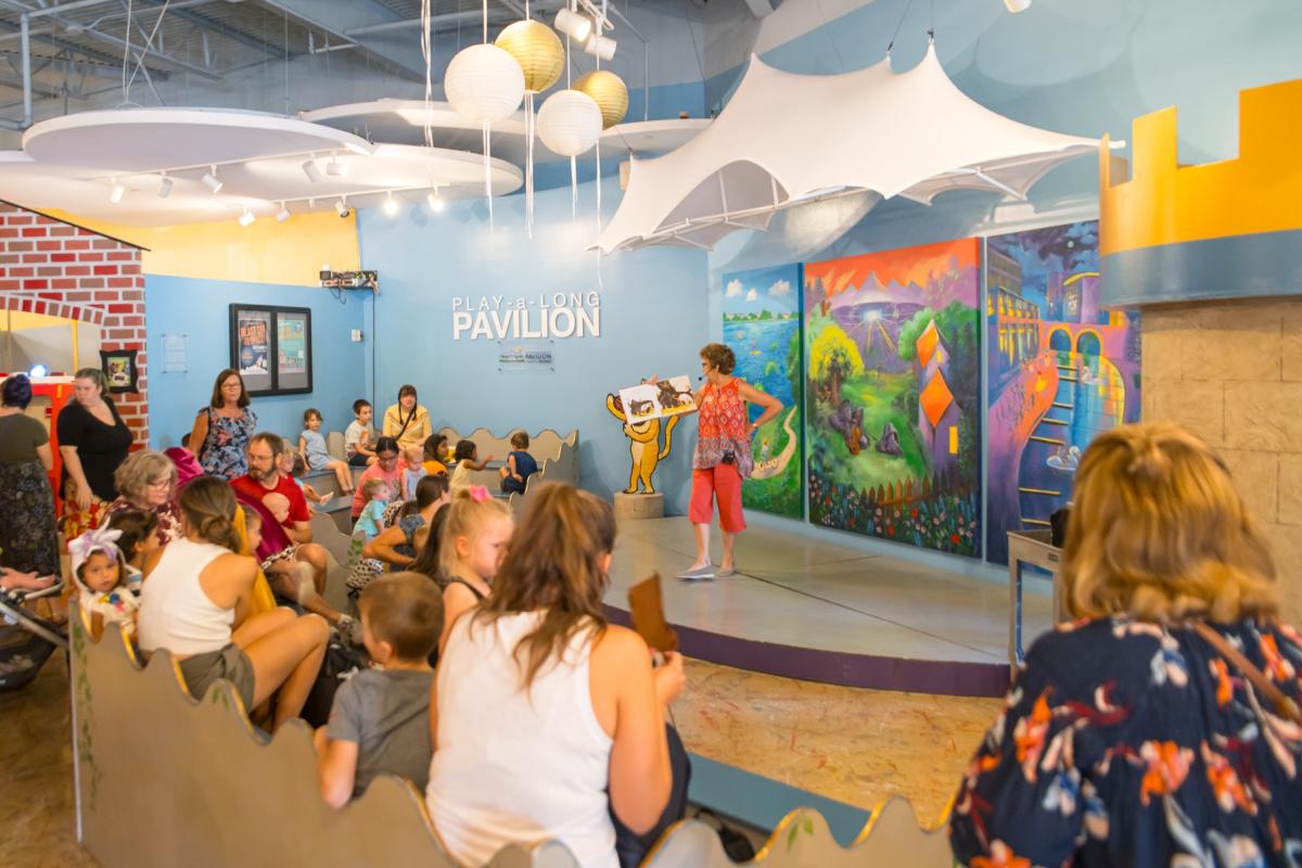 Inside TWCM is a stage set up to look like The Cynthia Woods Mitchell Pavilion, complete with tent awning and a tri-paneled mural with iconic landmarks around The Woodlands. The audience is full of families with young kids, all listening to a woman reading a children's book on the stage.