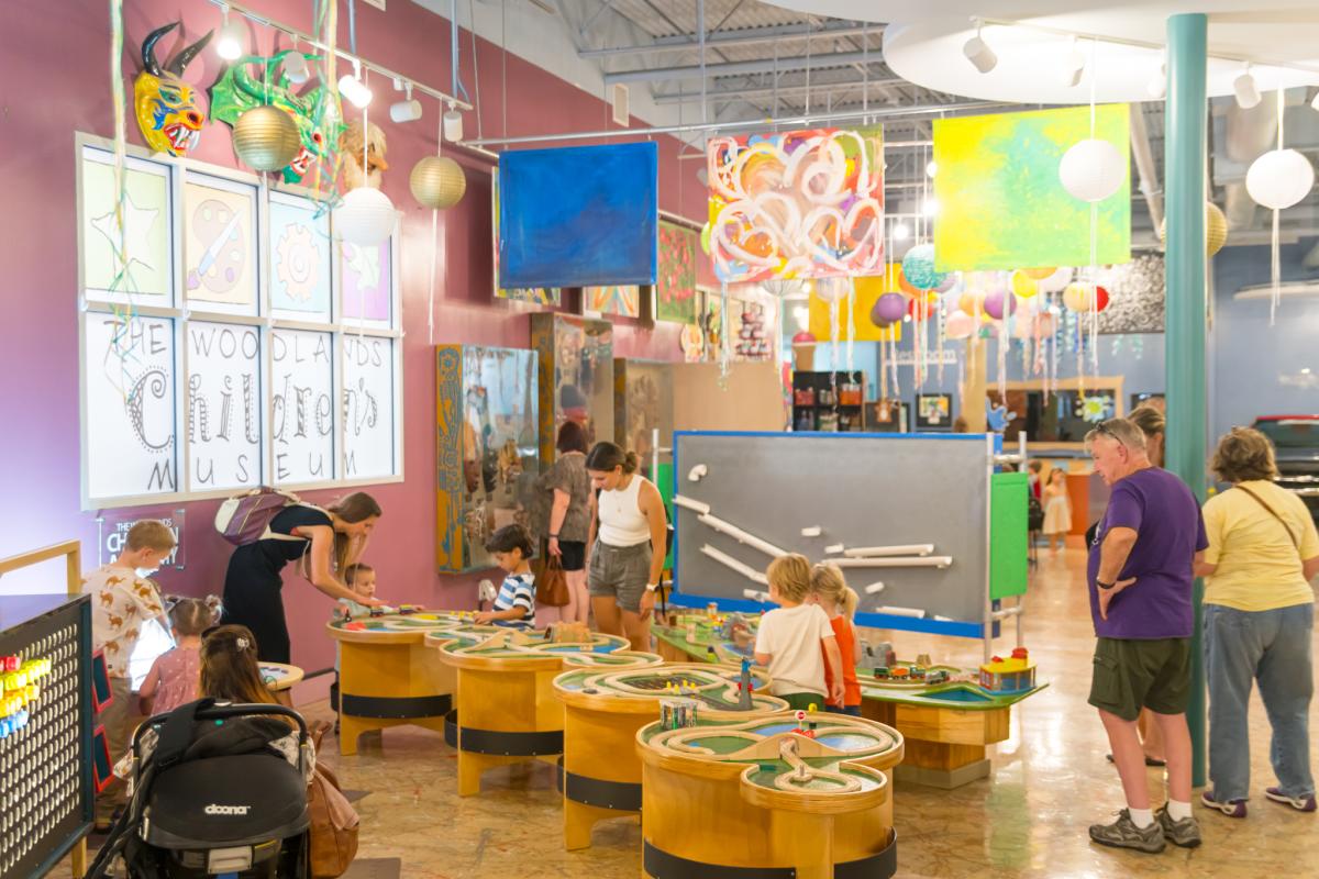 The interior of The Woodlands Children's Museum. The walls are painted purple and decorated with children's artwork. Light displays look like clouds and paper lanterns. There are exhibits with toy trains, a farmer's market, ships on the ocean, and more.