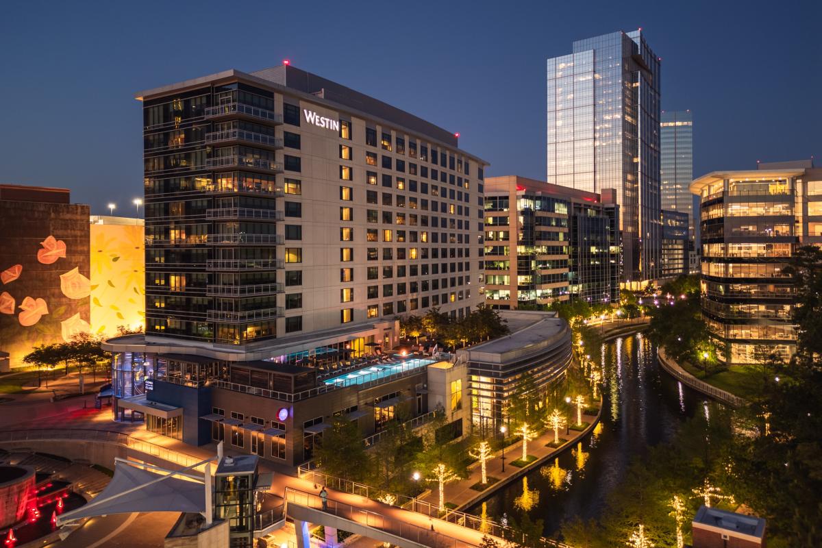 A drone shot of The Westin and The Woodlands Towers at dusk. Surrounding buildings and trees along The Waterway glow with soft golden lights.