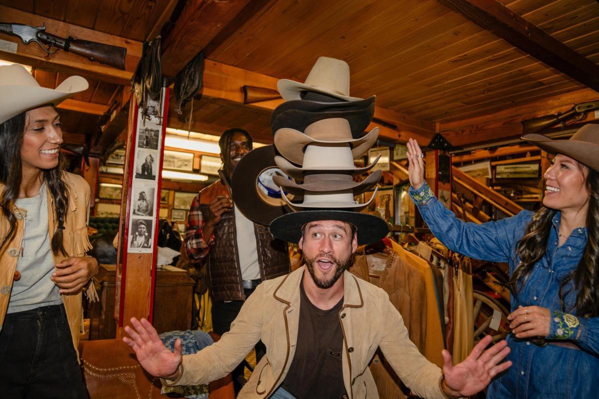 Four friends try on cowboy hats inside a rustic Western shop, with one man balancing a tall stack of hats on his head.
