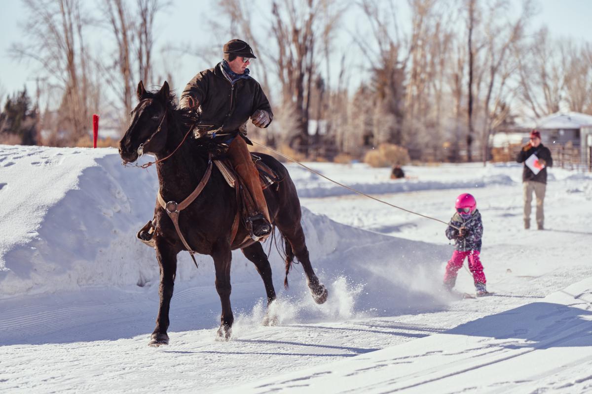 Little girl is competing in skijoring winter event in Pinedale, Wyoming, being pulled on a horse and rider on skis.
