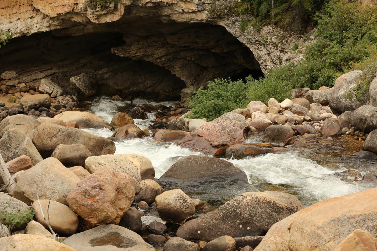 The Popo Agie River disappears beneath a limestone cliff at The Sinks in Sinks Canyon State Park near Lander, Wyoming. Whitewater flows around large boulders at the entrance to the underground cavern, showcasing one of the state's most unique natural geological features.