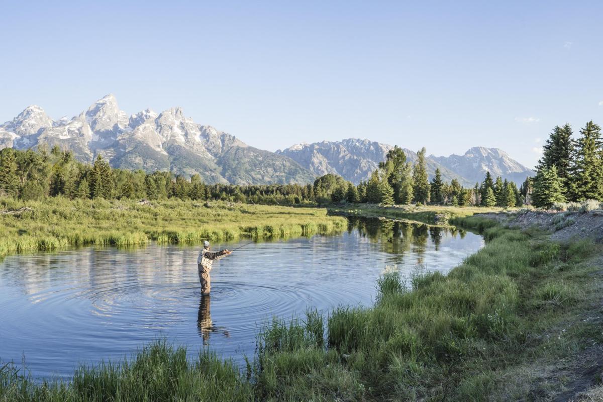 Man fly fishing in the middle of a river with mountians in the background.