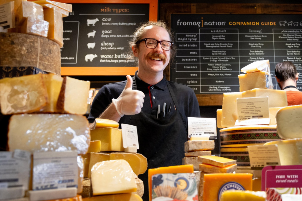 Cheesemonger at Fromagination counter surrounded by cheese giving a thumbs up