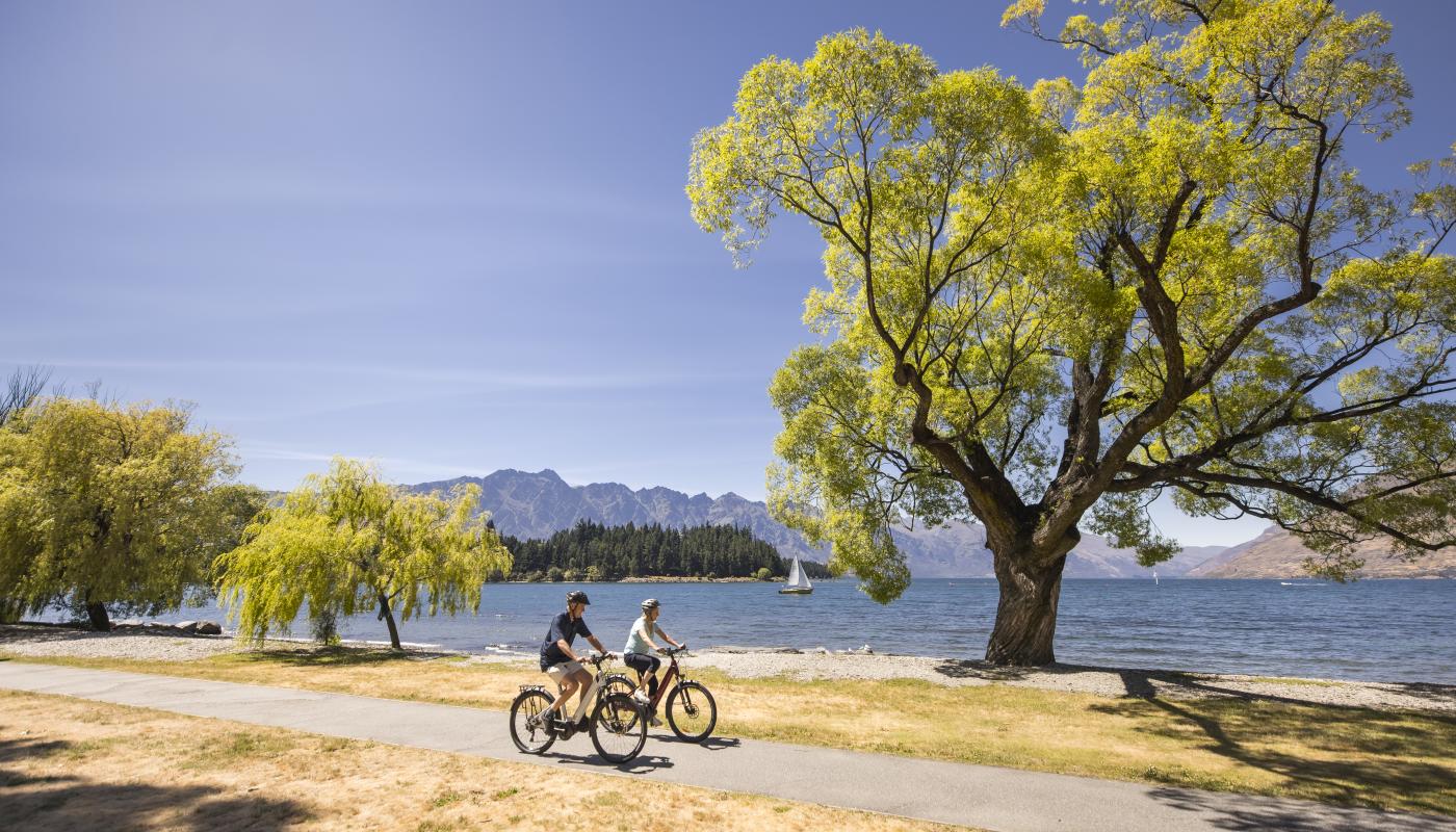 A couple bike along Queenstown Waterfront with the Remarkables mountain range in the background on a sunny day