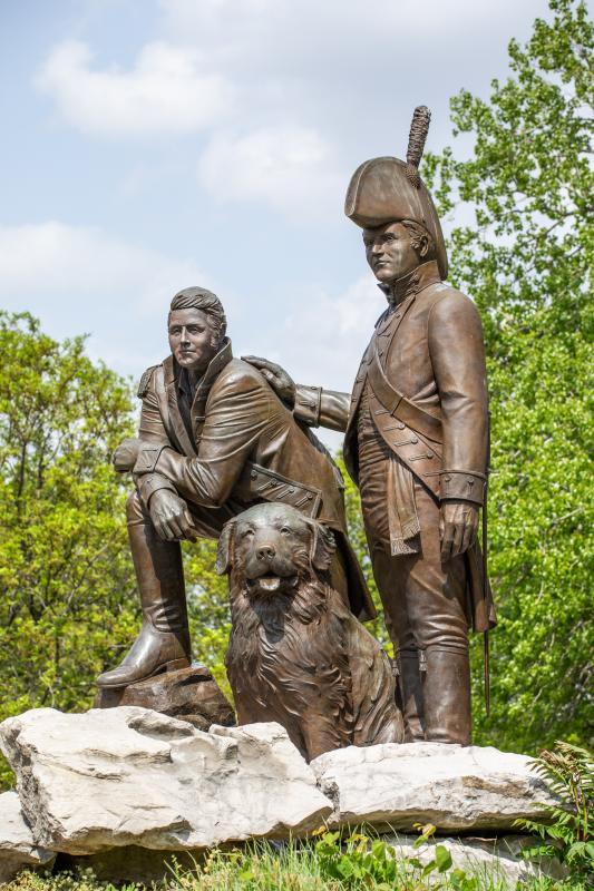 A larger-than-life statue of Lewis & Clark and their Newfoundland Dog, Seaman surrounded by trees in Frontier Park in St. Charles, Missouri.