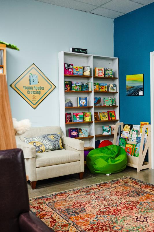 A reading nook for kids at Village Books, featuring tall and short bookshelves full of picture books, a sofa chair, a beanbag, a reading rug, and colorful animal plushies.
