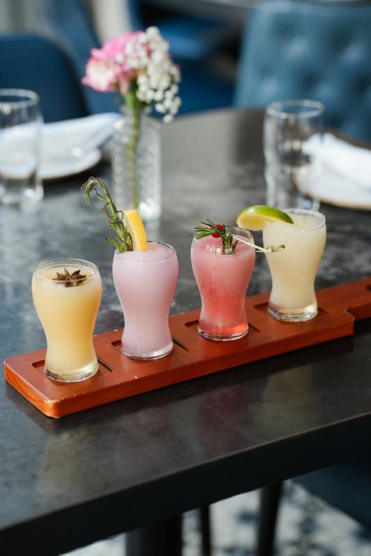 A flight of four frozen cocktails sit on a tray on the edge of a State Fare patio table. One is yellow with an anise seed pod floating on top; the second is pale purple with a lemon slice and herb sprig; the third is light red with a cranberry and sprig of rosemary; the last is pale yellow with a lime wedge.