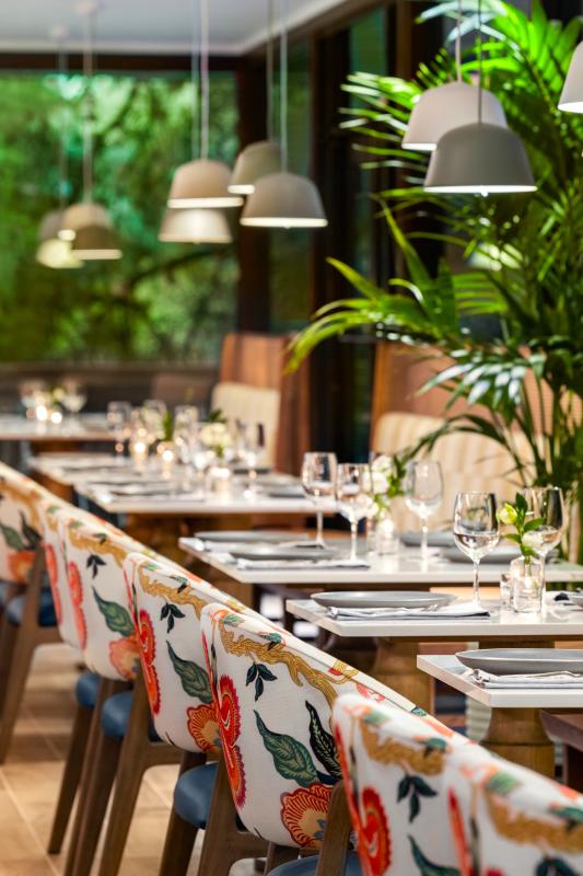 The interior of Harrison's features a row of two-person tables set with silver and white plateware. One side of the tables share a long booth, the other feature individual floral-cushioned chairs. Simple lights hang over every table, green foliage accents the room and out the windows.