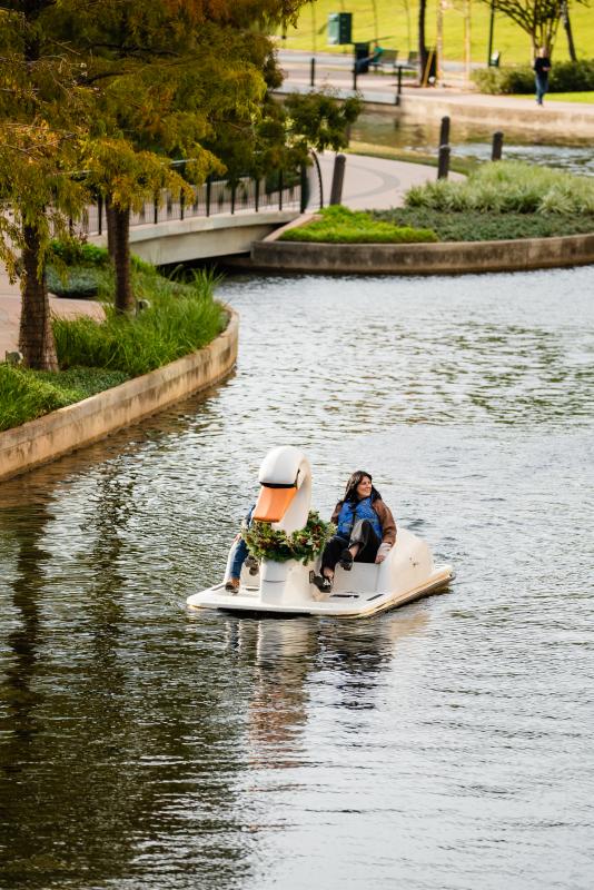 A couple paddles a swan boat down The Waterway (toward the camera). Only the woman is visible; she has long dark hair, a brown shirt, and black pants. She's also wearing a bright blue life jacket. The scenery behind them features a footbridge and fall foliage.