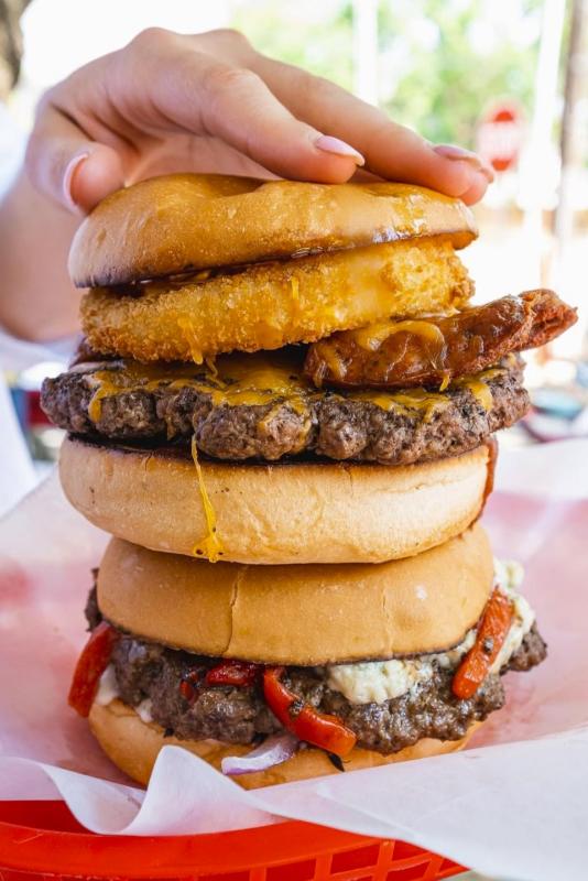 A woman's hand rests on top of one burger (onion rings and cheddar) stacked on another burger (grilled onion, pepper, and blue cheese).