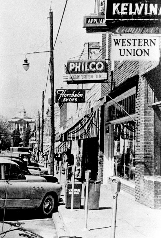 Mid-1950s Main Street sidewalk in Sylva, NC, with Philco, Florsheim Shoes, Western Union and Kelvinator signs, parking meters and the courthouse beyond