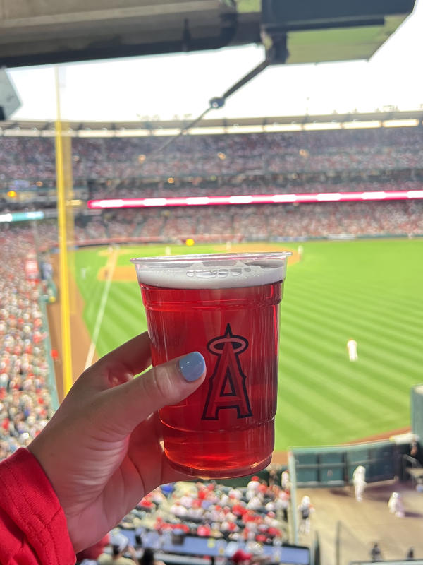 Image of a red beverage in a clear cup with an "A" logo. The cup is held in front of a green baseball field.