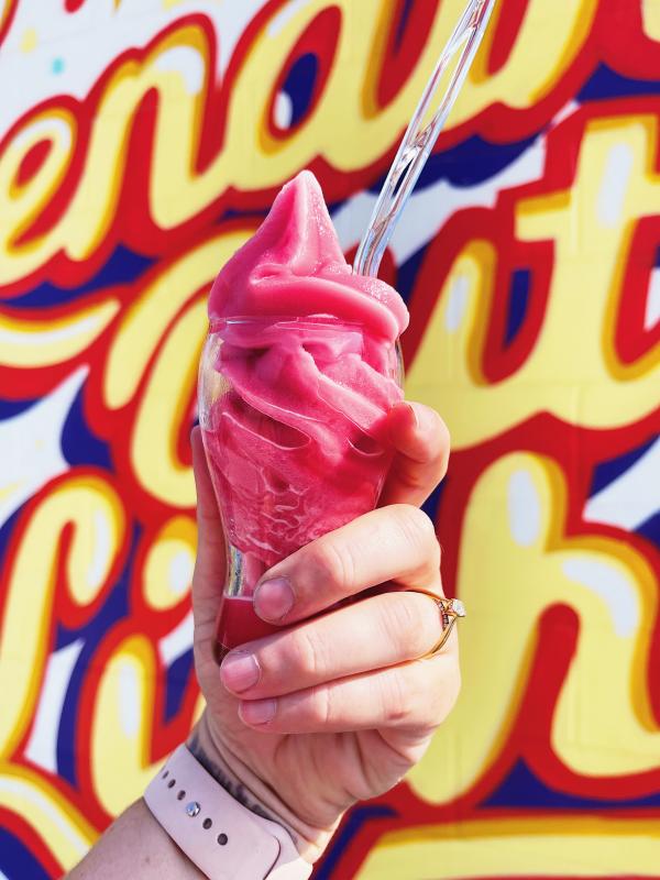 Image of a pink slushie drink, held upward. In the background red, yellow, and blue writing can be seen against a white wall.