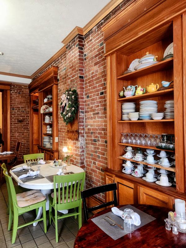 Cozy restaurant interior featuring a round table with green chairs, exposed brick walls, and a wooden shelf displaying fine china and glassware.
