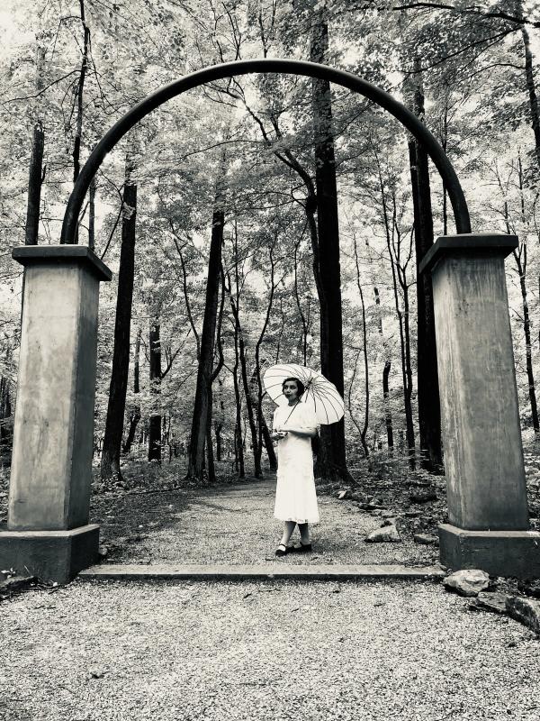 Historic black-and-white photograph recreation at Rose Island in Charlestown State Park, featuring a woman in a vintage white dress holding a parasol beneath the park’s iconic archway surrounded by wooded trails.