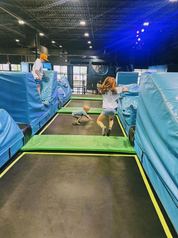 Children jump and play across a row of indoor trampolines surrounded by bright blue padded walls at Flow Trampoline Park in Huntsville, Alabama. Colorful overhead lights illuminate the activity area.