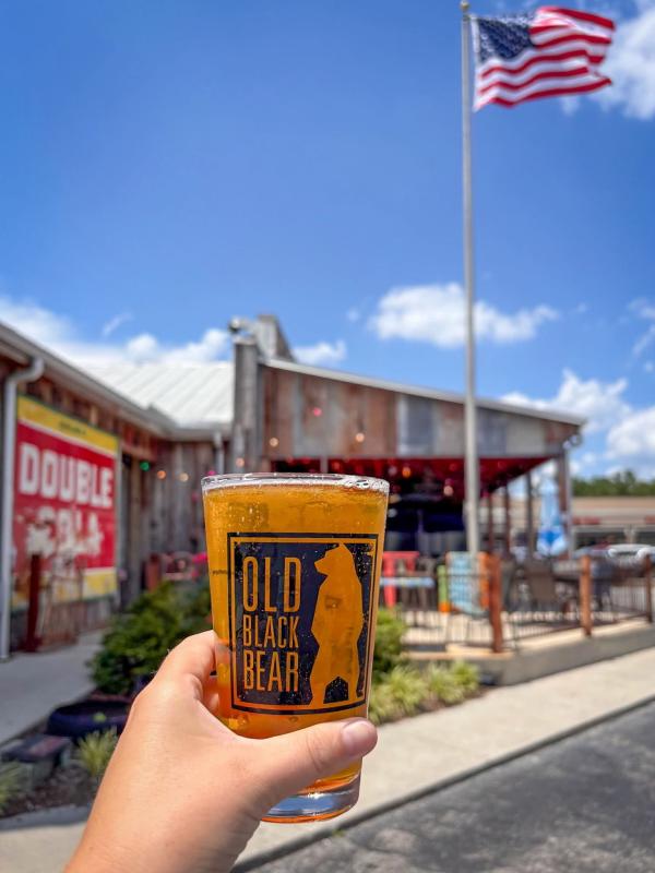Hand holding a glass of beer outside Old Black Bear Brewing Company, with an American flag and outdoor patio in the background.