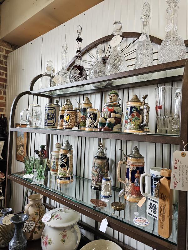 Display shelves inside Railroad Station Antiques filled with vintage collectibles, including ornate German beer steins, crystal decanters, glassware, ceramic pitchers, and decorative kitchenware. Many items have price tags, showcasing the shop's wide selection of antiques and unique treasures.