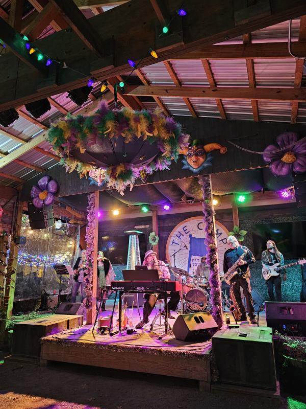 A live band performs on the outdoor stage at The Camp beneath colorful Mardi Gras decorations, string lights, and vibrant stage lighting. Musicians play keyboards, saxophone, bass, and drums as guests enjoy live music in the lively open-air venue.
