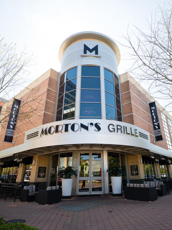 The exterior of Morton's Grille. It occupies the corner of a red brick building with a windowed tower facing the street. The signage is in bold black letters on a sleek metal face.
