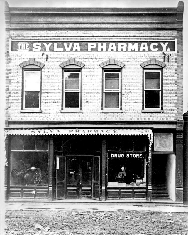 Two-story brick front of The Sylva Pharmacy drug store on unpaved Main Street in Sylva, NC, in the early 1920s, with an awning and open doors