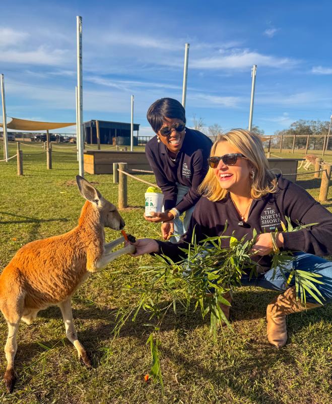 Zondra White Jones and Katie Guasco crouch in a grassy enclosure at Global Wildlife Center, smiling as they hand-feed a joey (young kangaroo) carrots and leafy greens.