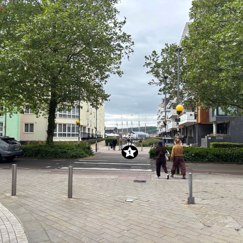 Millennium Promenade Social Story Picture 2 - A photo of a paved area leading to a zebra crossing over a road, there are three silver bollards before the crossing, two people ahead and some large trees on the other side of the road.
