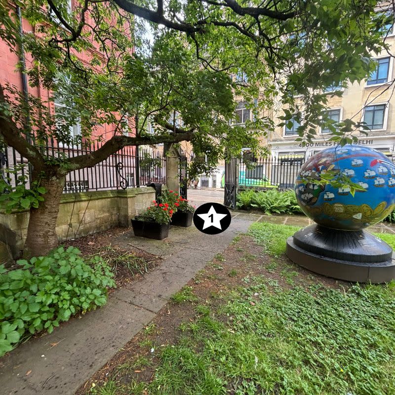 St Stephens Church Gardens Social Story Picture 10 - A photo of a narrow path with a tree and a flower bed on the left side. On the right of the path is a lawn area with a big blue ball sculpture on. There is a gate at the end of the path leading out of the garden.