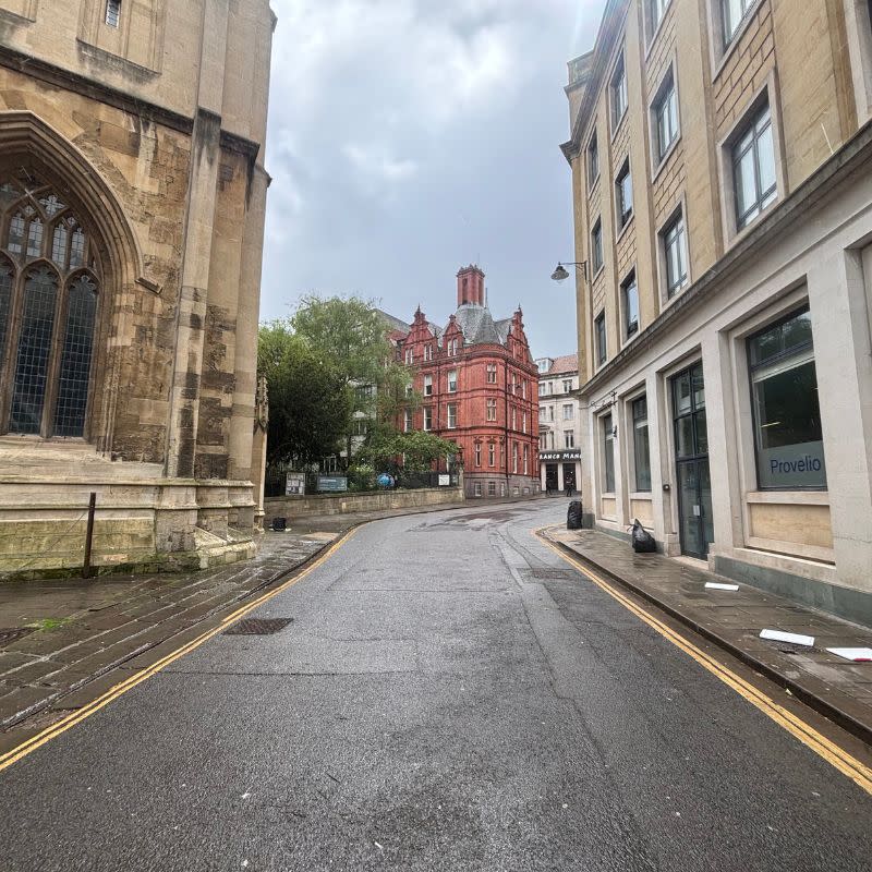 St Stephens Church Gardens Social Story Picture 1 - A photo of an empty street with cream coloured buildings on each side. In the distance there is a churchyard with mature trees and a red brick tall building.