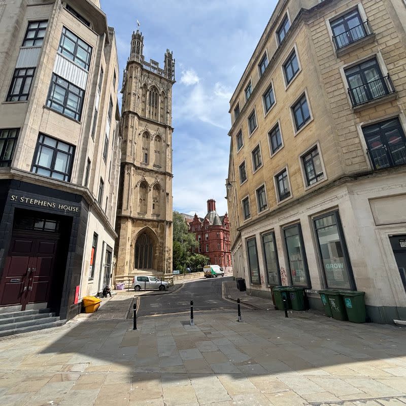 St Stephens Church Gardens Social Story 1 - This picture shows wide path with tall buildings on either side, the path is leading up to a narrow empty road with a church on the left hand side. Up ahead is a tall red brick building.